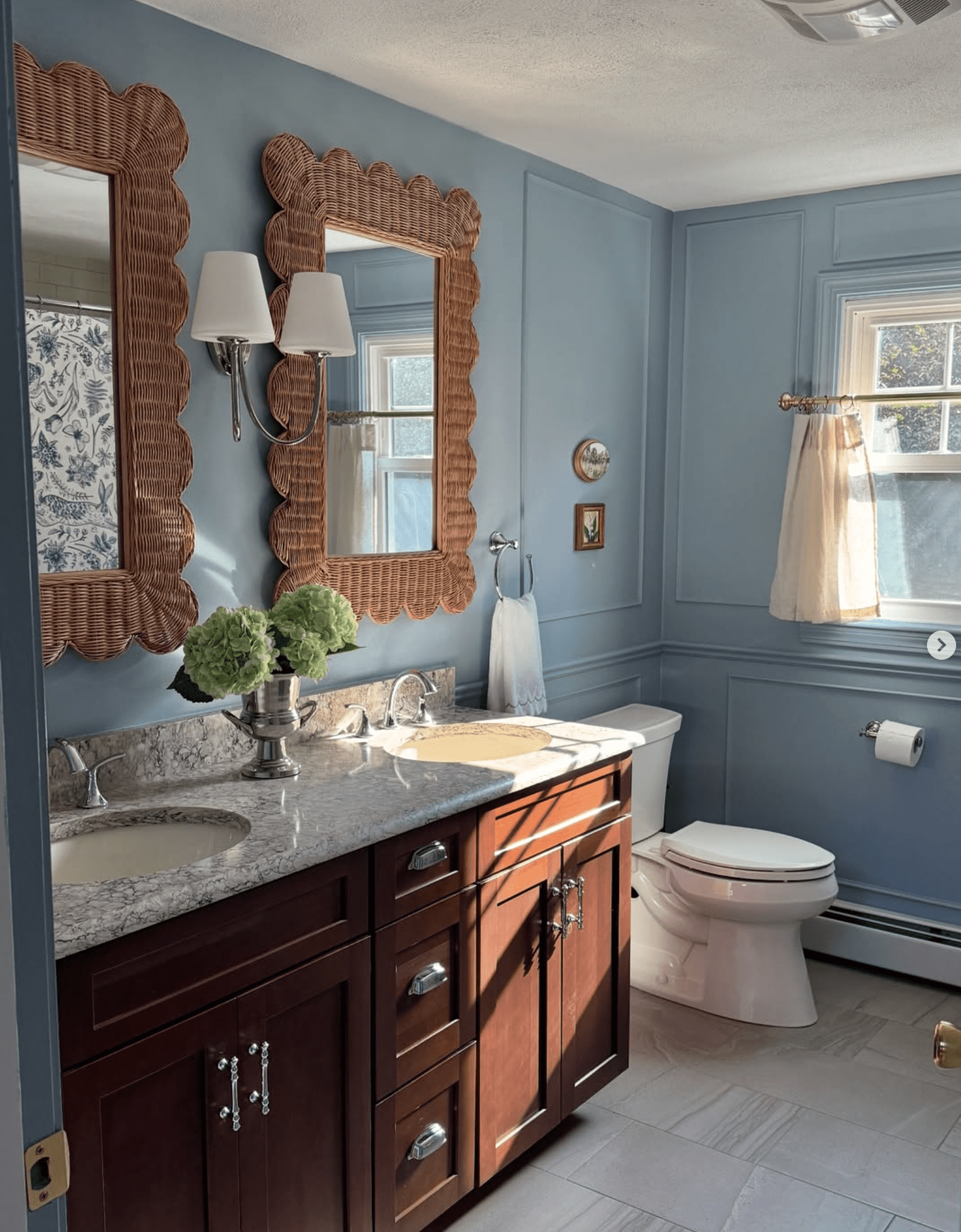 Grandmillennial-style bathroom with blue paneled walls, wicker scalloped mirrors, marble double vanity with dark wood cabinets, silver fixtures, and soft neutral tile flooring — budget-friendly renovation in a Rhode Island home.