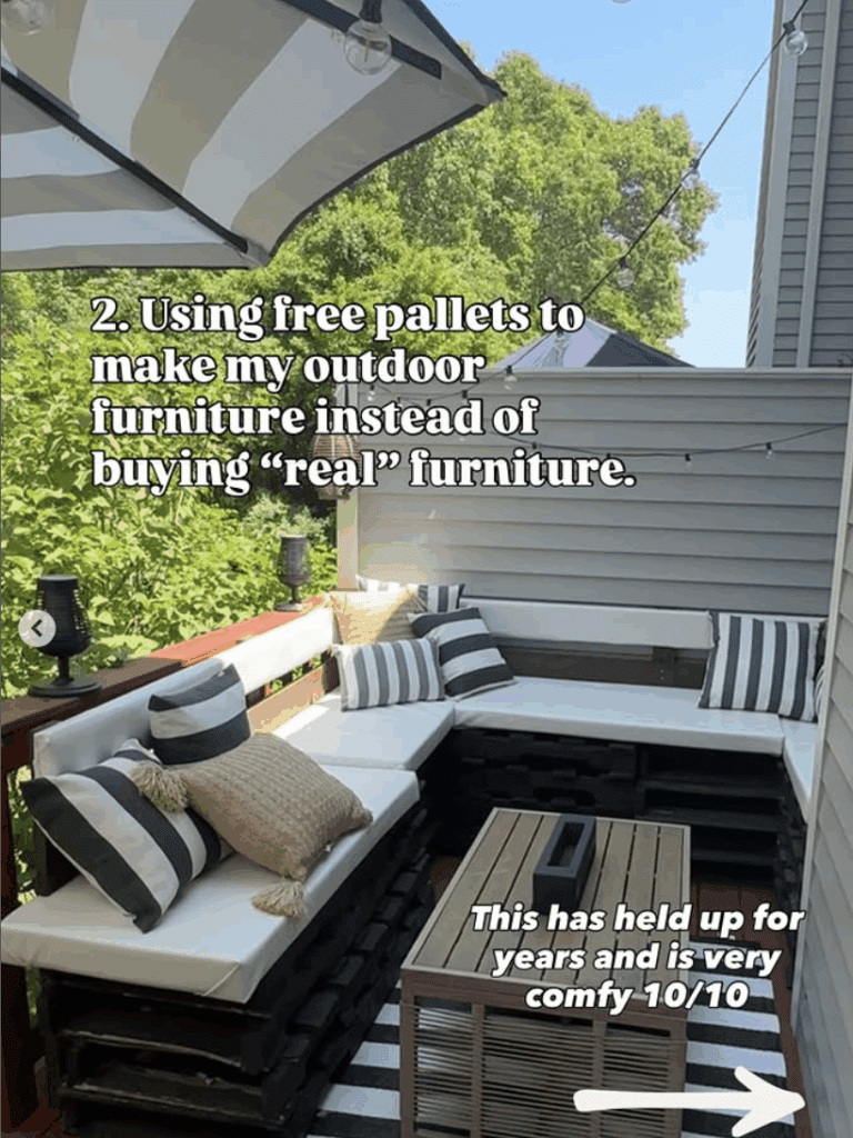 DIY outdoor sectional sofa made from repurposed wood pallets, painted black, with white cushions and black-and-white striped throw pillows on a small deck with a striped outdoor rug and large umbrella. Budget-friendly, cozy, and sustainable patio furniture idea.