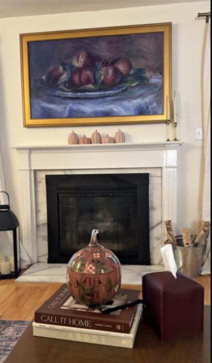 Cozy traditional fall living room with marble fireplace, gold-framed fruit painting, ceramic pumpkins, and fall accents on coffee table with seasonal pumpkin decor.