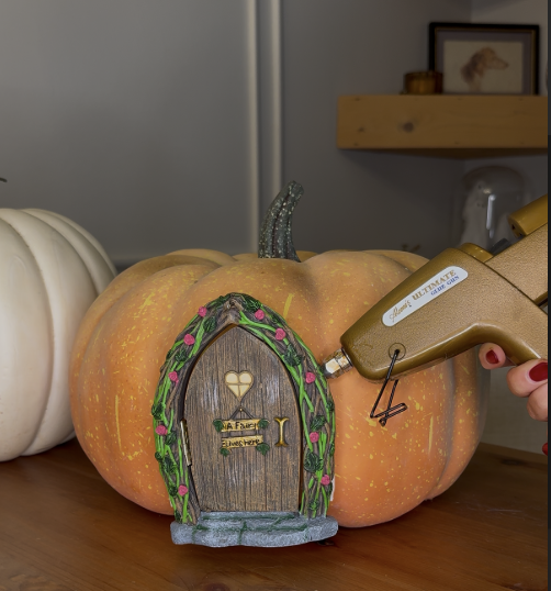 Applying hot glue around Tree Poetry fairy door on faux pumpkin to complete whimsical fall DIY pumpkin decoration.