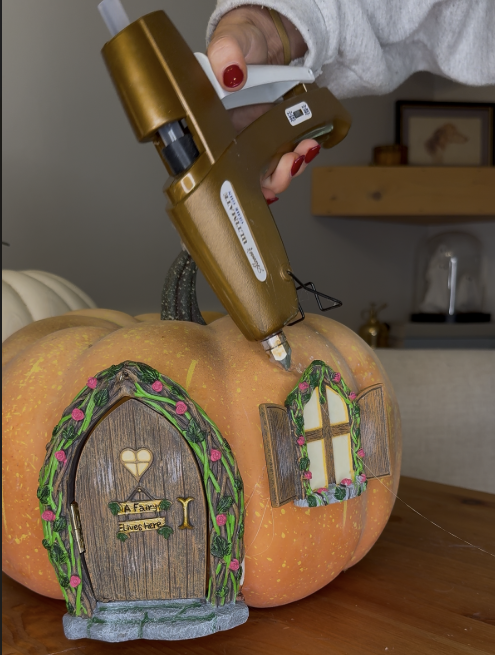 Using hot glue gun to secure Tree Poetry fairy window onto faux pumpkin while creating DIY fairy door pumpkin project.