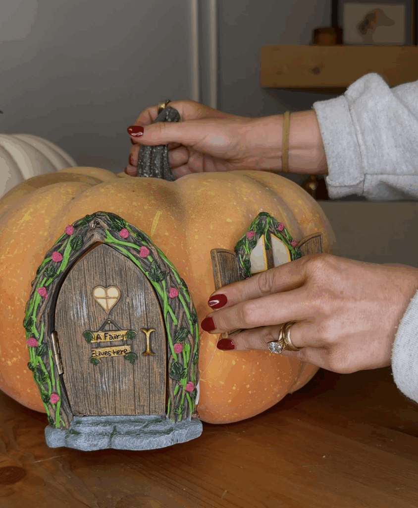 Hands securing fairy window onto faux pumpkin next to fairy door using hot glue for easy DIY pumpkin craft.