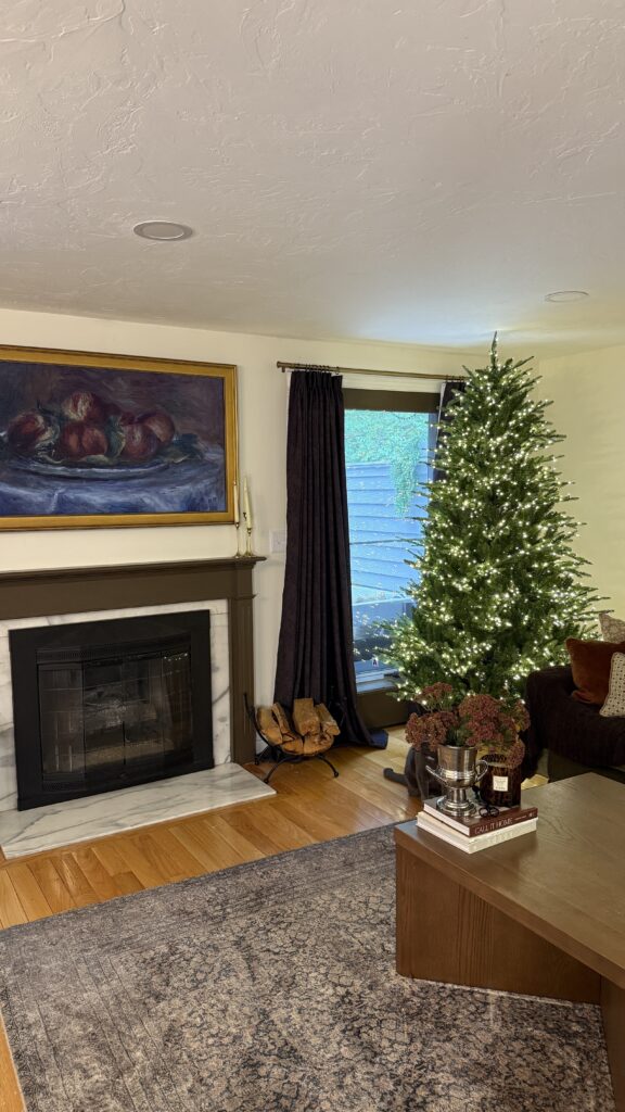 7.5 ft Grand Duchess twinkling Christmas tree with warm white LED lights placed by a window in a cozy traditional living room with fireplace and wood floors.