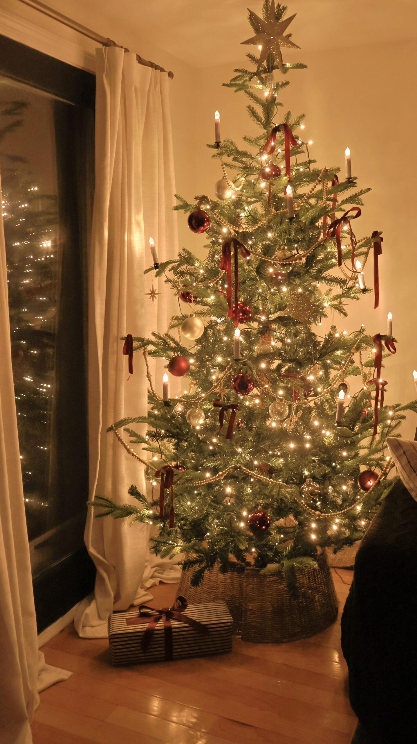 Cozy Christmas tree decorated with red velvet ribbons, gold garland, glass ornaments, and warm candle lights, styled in a woven basket base beside white curtains.