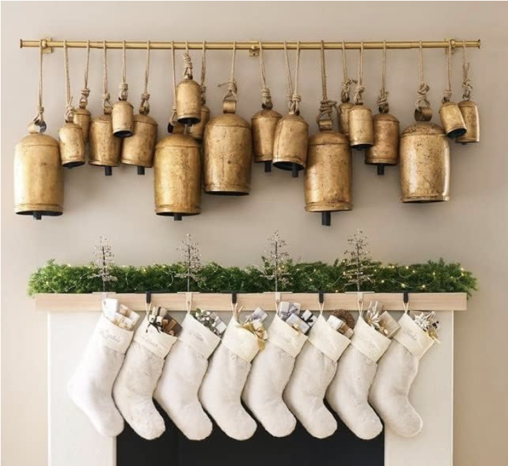 Elegant brass bell garland hanging over a mantel decorated with greenery and white Christmas stockings. A timeless, cozy holiday look featuring rustic metallics and natural textures for traditional and classic Christmas home styling.