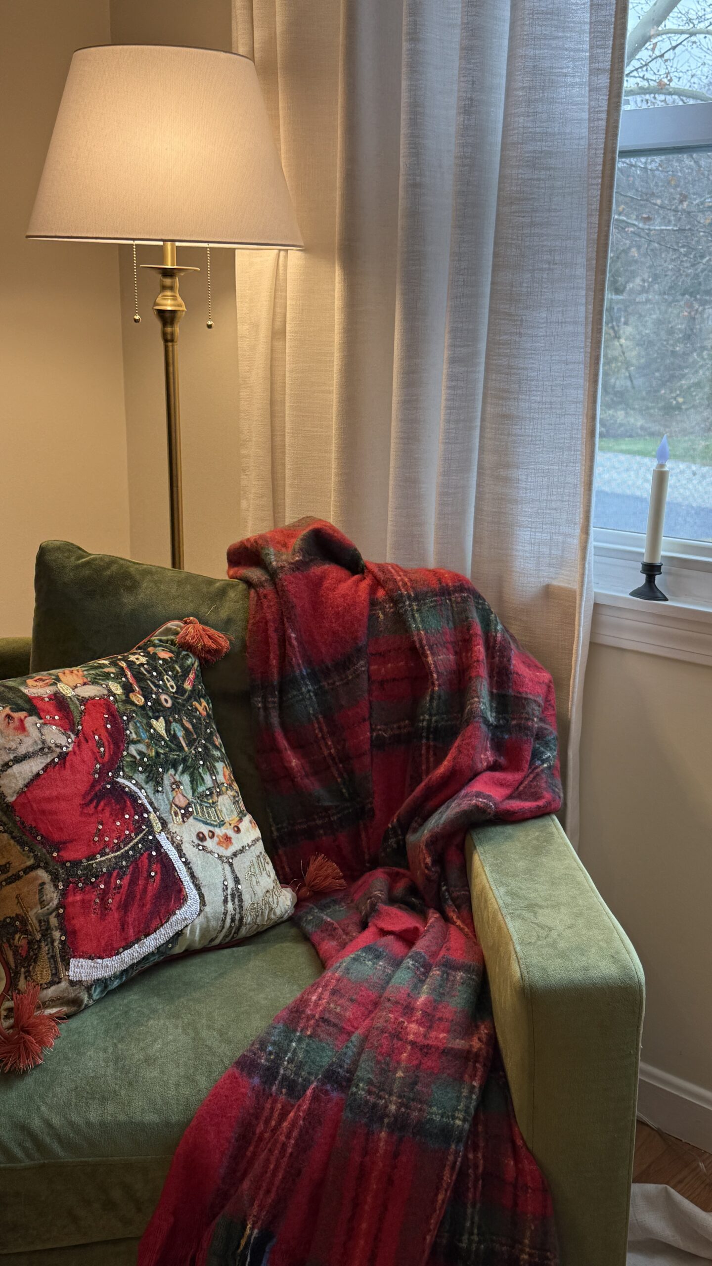Cozy Christmas living room corner featuring a brass floor lamp from wayfair, cream linen curtains, and a green velvet armchair draped with a red tartan plaid throw blanket. A vintage-style Santa pillow adds nostalgic holiday charm, with soft window candlelight creating a warm, festive glow.