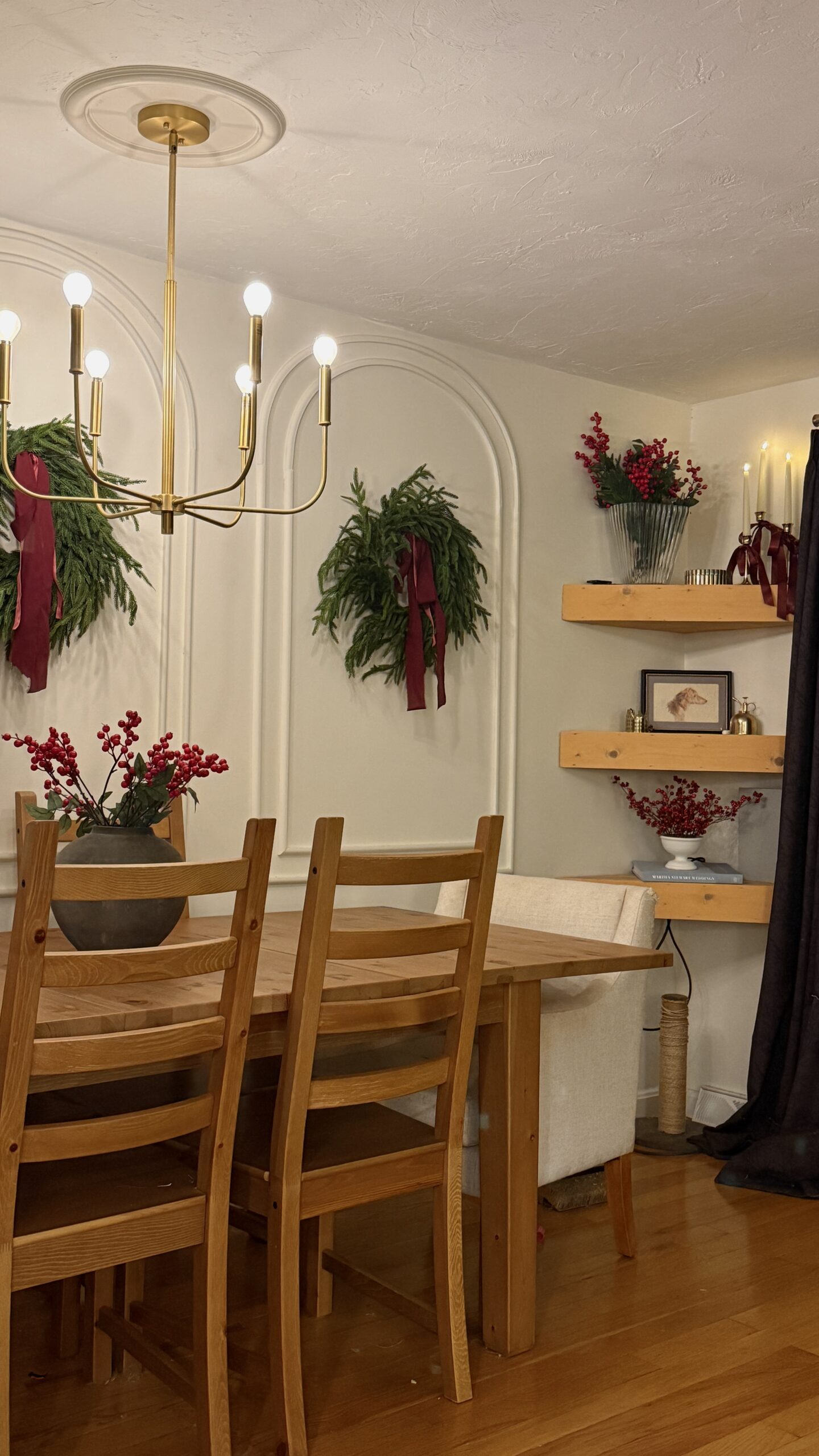 Natural-wood dining room decorated for Christmas with simple greenery wreaths, red velvet ribbon, a brass chandelier, floating shelves styled with berries and candles, and a cozy, classic holiday look. If you want a meta description or a blog-post intro to go with this, just let me know!
