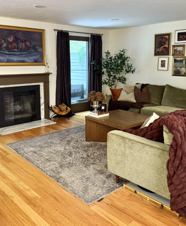 Neutral living room before Christmas decorating, featuring a green sectional sofa, wood coffee table, fireplace, and layered cozy textures. The space has warm lighting, natural wood floors, and everyday decor before being styled for the holidays.