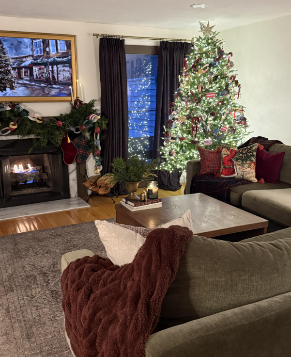 Home office decorated for Christmas with a lit tree trimmed in ribbons and paper chains, a burl wood desk, Santa mug, and a small electric fireplace heater. Diploma wall frames and woven baskets add character to the cozy, festive workspace.